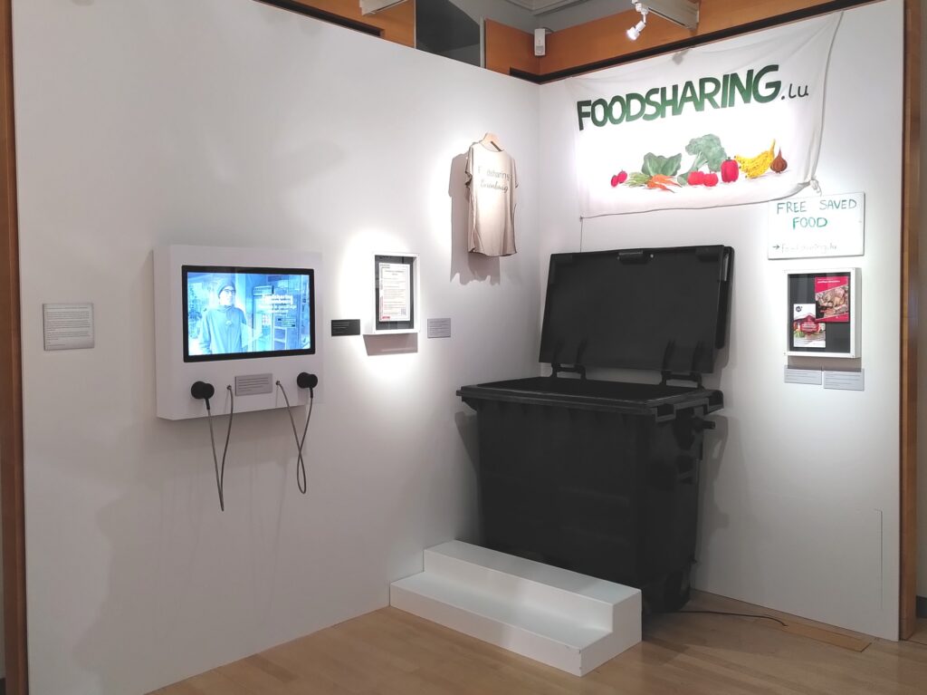 Wall with a screen with a video about Foodsharing Luxembourg, a framed flyer, a t-shirt with the text Foodsharing Luxembourg, a banner with the text foodsharing.lu, a sign reading Free Saved Food, a big black bin and two other flyers